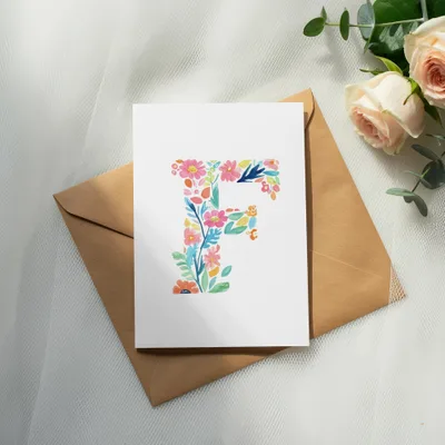 Watercolor letter F with floral accents on a card atop a kraft envelope.