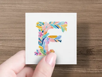 Watercolor floral letter 'F' on a white card held by a hand.