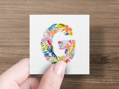Floral letter G on a card held by a hand.