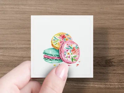 Three watercolor macarons with floral designs on a white card held by a hand.