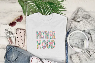 White t-shirt with "Motherhood" text in floral design, laid flat with clothing and accessories.