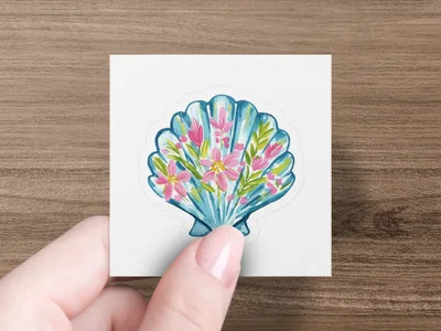 Blue seashell sticker with pink and green floral design held by a hand.