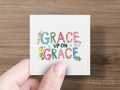 Hand holding square card with colorful watercolor text "GRACE UPON GRACE."