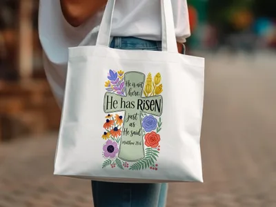 White tote bag with floral cross graphic and "He has risen" text.