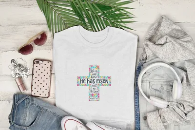 White t-shirt with a floral cross and "He is not here, He has risen" text.