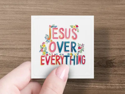 "JESUS OVER EVERYTHING" text with floral accents on a white card held by a hand.