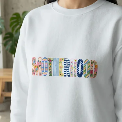 White sweatshirt with "MOTHERHOOD" in colorful, patterned block letters.