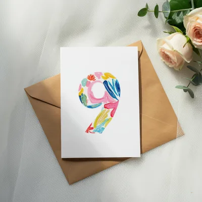 Watercolor number nine with floral patterns on a card, next to an envelope and roses.