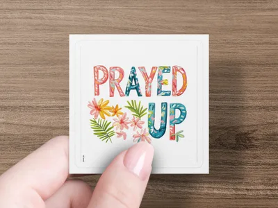 Square coaster with "PRAYED UP" text and floral accents held by fingers.