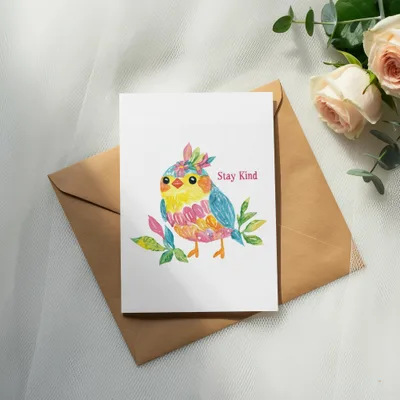 Watercolor bird with "Stay Kind" text on card, resting on brown envelope.