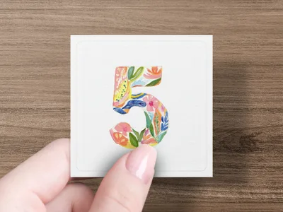 A hand holds a square card with a colorful floral number 5 on a wooden background.