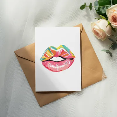 Watercolor lips art on card atop envelope with roses and eucalyptus.