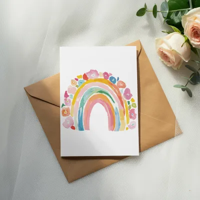 Watercolor rainbow art on a card atop a brown envelope, with roses and eucalyptus.
