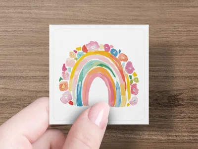 Hand holding a square card with a watercolor rainbow and floral border.