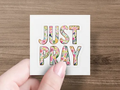 Floral "JUST PRAY" text on a sticker held by a person's hand.
