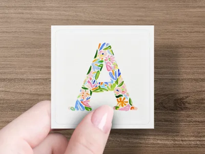 Letter A in floral watercolor design held by a person's fingers.