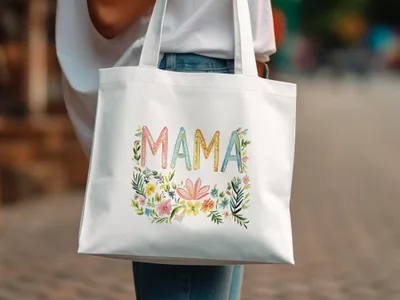 White canvas tote bag with colorful "MAMA" text surrounded by floral illustrations.