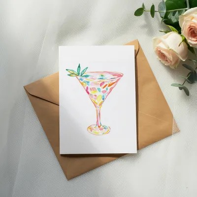 Watercolor cocktail glass with colorful splashes on white cardstock atop a brown envelope.