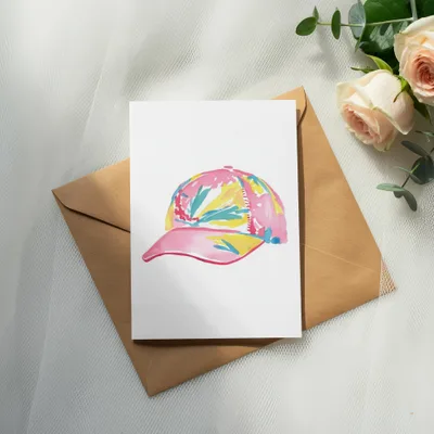 Pink baseball cap with colorful abstract pattern on white card over brown envelope.