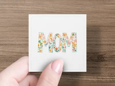 Floral "Mom" text on a square card held by a hand.