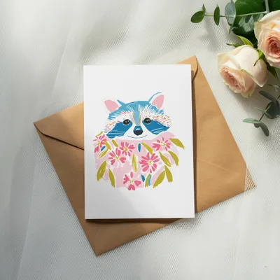Card with raccoon illustration wearing a floral wreath, on an envelope.