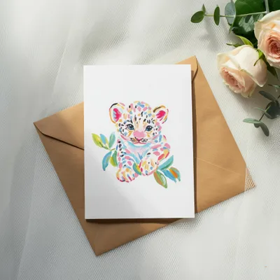 Watercolor painting of a colorful baby leopard cub on a card atop a brown envelope.