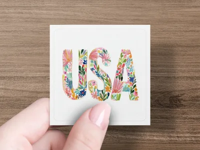 Floral "USA" lettering on a white square card held by a hand.