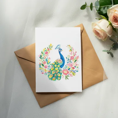 Watercolor peacock with floral wreath on a card atop a brown envelope.