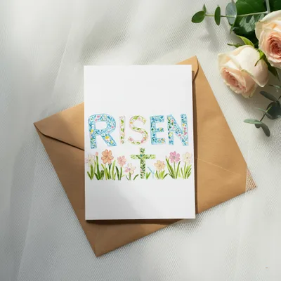 Card with "RISEN" and a floral cross on a kraft envelope.
