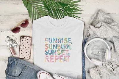 White t-shirt with "Sunrise, Sunburn, Sunset, Repeat" graphic, laid flat with clothing and sunglasses.