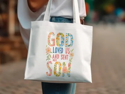 White tote bag with colorful floral lettering that reads "God Loved Us And Sent His Son."