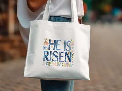 White tote bag with "HE IS RISEN" in blue lettering and floral accents.