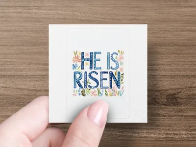 "He is Risen" text with floral accents on a sticker held by a hand.