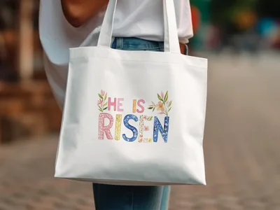 White tote bag with "He is Risen" text in colorful floral letters.