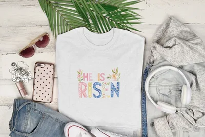 White t-shirt with "He is Risen" text, surrounded by jeans and headphones.