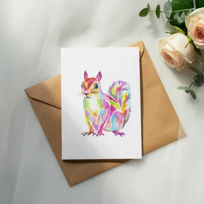 Colorful watercolor squirrel illustration on a card, next to an envelope and roses.