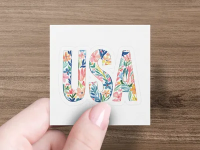 Floral letters spell "USA" on a white sticker held by a hand.