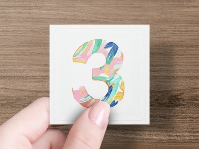 Hand holding square card with colorful abstract number three painted on it.