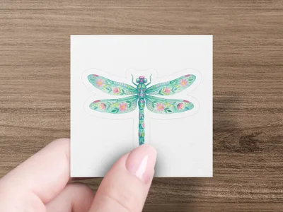 Dragonfly sticker with floral wing patterns on white paper held by a hand.