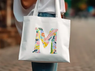 White tote bag with a large colorful floral letter "M" design.