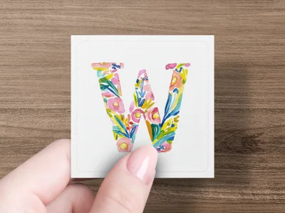 Hand holding a square card with a floral letter W.