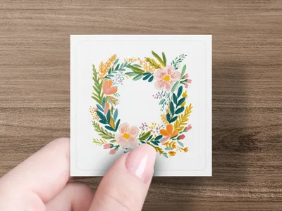 Square card with floral wreath illustration held by a person's hand.