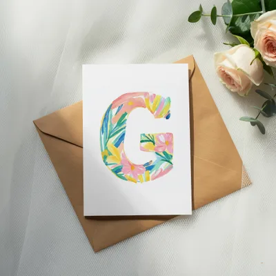 Watercolor letter G with flowers and leaves on cardstock, next to roses.