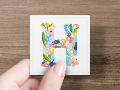 Hand holding a square card with a watercolor letter "H" design.