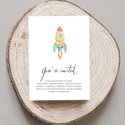 Card with "You're invited" text and a rocket illustration, on wood slice.