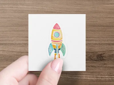 Colorful rocket ship sticker with star accents on a white background held by a hand.