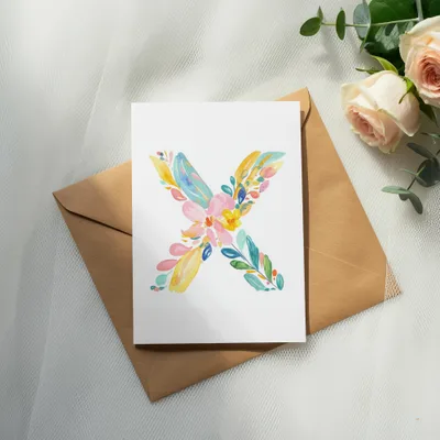 Watercolor letter X made of colorful flowers on a card atop an envelope.